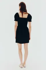 Vestido corto negro de popelina de algodón con escote cuadrado y mangas cortas abullonadas con elástico. El cuerpo es ajustado con fruncido y la falda con vuelo. Tiene ribete de volado en escote y puños.