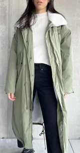 Campera larga color verde militar con cuello de piel sintética blanca, cierre con botones a presión y cordón ajustable en la cintura.