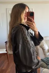 Chaqueta tipo bomber corta de cuero ovino color chocolate oscuro, forrada en su interior. Presenta detalles de pasamanería en forma de cruz en cuero marrón desgastado en ambas mangas y botones de guampa.