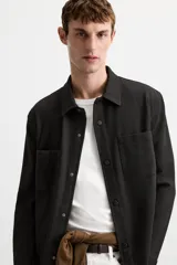 Sobrecamisa negra de tejido con elasticidad, cuello solapa y manga larga acabada en puño con botón a presión. Tiene bolsillos de plastrón en el pecho y cierre frontal con botones a presión.
