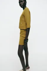 Chaqueta color mostaza de tejido de punto, con cuello redondo y manga tres cuartos. Tiene bolsillos delanteros de parche y cierre frontal con botones dorados.