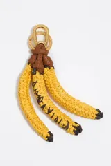 Llavero con dije que simula un racimo de tres bananas, hecho de rafia trenzada amarilla con detalles en negro que simulan las marcas de la fruta. La parte superior está sujeta con cuero marrón y un mosquetón metálico dorado.