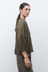 Blusa color verde oliva de manga 3/4 con volumen, cuello mao con abertura en V y botones. Presenta bordados y detalles de broderie calado en el canesú frontal y en las mangas. El cuerpo tiene un corte peplum con frunces.