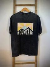 Remera negra con efecto gastado, corte amplio y estampado de montañas con la palabra 'Mountain' en blanco.