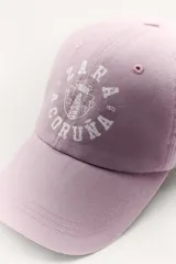Gorra color borgoña con bordado dorado de la marca Zara, con la inscripción "Zara" y "A Coruña 1975".