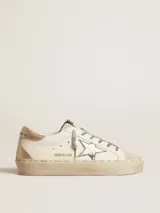 Championes de cuero Golden Goose modelo Hi Star, color blanco con detalles en dorado y plateado. Presentan una estrella de glitter plateado en el lateral, puntera de gamuza gris y talón en cuero dorado. La suela de plataforma incluye inscripciones de la marca y un acabado sutilmente desgastado.