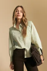 Camisa blanca de manga larga con cuello y botones frontales. Presenta detalles de volados o puntilla en los hombros y mangas abullonadas con puños anchos. La tela parece tener un sutil tejido a rayas verticales.