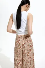 Pantalón de corte holgado en tejido dobby color marrón con estampado de hojas en color crema. Modelo de tiro alto con elástico en la parte posterior de la cintura y cierre oculto con corchete en un lado. Bolsillos laterales discretos y piernas amplias con pliegues adelante y atrás.