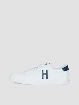 Zapatillas urbanas de corte bajo, color blanco, con cordones y suela de goma. Presentan un gran logo 'H' en color azul marino en el lateral y detalles en azul marino en el talón y la lengüeta.