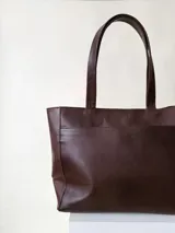 Cartera tipo tote color beige, hecha en cuero graneado con textura uniforme y suave al tacto. Tiene doble asa de hombro, un bolsillo frontal con broche imantado, un bolsillo trasero con cierre metálico y cierre principal también metálico. El interior está forrado y cuenta con tres bolsillos: uno con cierre, uno tipo plaqué para celular y un bolsillo grande tipo sobre con solapa y botón.