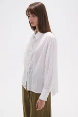 Camisa blanca de broderie con cuello camisero, cierre frontal de botones, mangas largas y corte holgado.