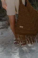 Bolso grande tejido a crochet con trapillo color marrón, con flecos en la parte inferior y etiqueta de cuero con el logo de la marca.
