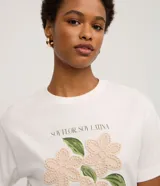 Remera blanca de algodón, con cuello redondo y manga corta. Presenta un estampado frontal con la frase "SOY FLOR. SOY LATINA" y un diseño floral con flores en relieve y hojas bordadas.