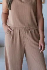 Pantalón ancho de tiro alto, color tostado, con finas rayas verticales blancas. Presenta cintura fruncida con elástico y una hilera de botones decorativos en el frente.