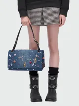 Bolso de hombro rectangular de denim azul, con solapa frontal decorada con múltiples charms metálicos y de esmalte, incluyendo cerezas, lazos, estrellas y candados. El bolso tiene herrajes dorados y correa de hombro de cuero negro.