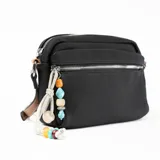 Bandolera negra de tela con correa larga ajustable en color beige. Tiene un bolsillo frontal con cierre y un adorno colgante de cuentas de colores.