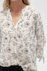 Blusa color beige con estampado de arabescos blancos, cuello mao, mangas 3/4 con lazos en los puños, cierre parcial con botones y bolsillos frontales.