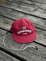 Gorra de béisbol color rojo vino con visera curva. Estampado frontal con un corazón y el texto "LOVE & PEACE" en blanco, y debajo "PP-ONE".