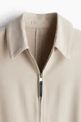 Campera color beige de tejido suave con cuello, cierre frontal, puños ajustables con botón a presión y bolsillos insertados al bies. Corte estándar y mangas forradas.