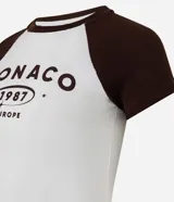 Remera ajustada de algodón con mangas cortas estilo raglán en color marrón oscuro y cuerpo blanco. Presenta un estampado central con el texto "MONACO 1987 EUROPE" en color marrón.