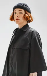 Camisa negra de manga corta, corte oversize, confeccionada en poplin 100% algodón. Presenta dos bolsillos delanteros, uno con solapa y otro con ojal y botón.