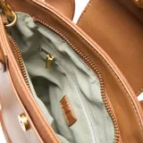 Cartera tipo tote bag de material símil cuero color habano, con cuerpo principal de tela texturada en tono similar. Posee doble asa corta, correa larga desmontable y regulable, cierre superior metálico y bolsillo interno con cierre. Presenta un bolsillo frontal con solapa de cuero y herrajes dorados.