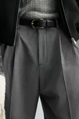 Pantalón gris de corte ancho con cintura elástica, pliegues frontales, bolsillos laterales y traseros de vivo. Incluye detalle de hebilla metálica en la trabilla y cierre frontal con cremallera y botón.