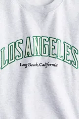 Buzo gris con cuello redondo, mangas largas y puños acanalados. Estampado frontal con el texto "Los Angeles Long Beach, California" en color verde.