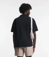 Camisa negra de sarga, corte boxy, con cuello inglés, manga corta, abotonadura frontal y bolsillo en el pecho.