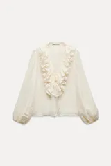 Blusa color crudo confeccionada en hilatura con mezcla de lyocell. Escote pico ajustable con cordones, manga larga con volumen acabada en elástico y detalle de volantes en el mismo tejido.