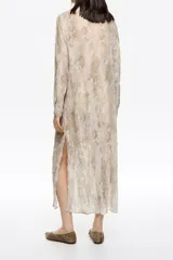 Vestido largo de corte recto confeccionado en tejido plisado con estampado de piel de serpiente en tonos beige y marrón. Presenta botones frontales, cuello camisero, mangas largas con puño y botón, y maxilazos en el escote delantero.