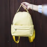 Mochila color mostaza de símil cuero con bolsillo frontal con cierre y broche, apertura superior con cierres metálicos, bolsillos laterales y asa de mano. Incluye bolsillos internos y correas regulables para colgar en la espalda.