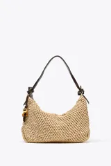 Bolso tipo shopper color beige, con cuerpo trenzado de rafia y asa de hombro de cuero marrón. Tiene un charm colgante en un lateral y cierre con cremallera.