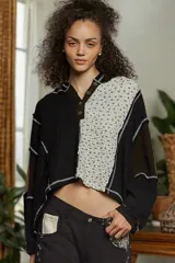 Buzo cropped de manga larga, color negro, con cuello tipo polo y cierre parcial de botones. Presenta un diseño de patchwork con paneles de tejido acanalado negro y un panel central de tela estampada con microflores blancas, todo unido con costuras expuestas en hilo blanco.