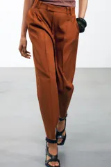 Pantalón color marrón confeccionado en hilatura con mezcla de lyocell y algodón. Tiro medio, cintura con trabillas, bolsillos laterales, pinzas en delantero y raya planchada. Cierre frontal con cremallera, botón y ganchos metálicos.