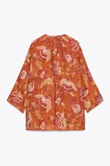 Camisa arrugada de ramio color naranja con estampado floral en tonos rosados y blancos. Tiene cuello redondo con escote pico ajustable con lazadas, mangas 3/4 y bajo asimétrico con aberturas laterales.
