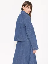 Campera de denim azul con textura en relieve, corte recto, cuello camisero, cierre frontal con botones blancos, bolsillos delanteros y mangas largas con pinzas que generan volumen.