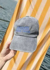 Gorro de béisbol color gris desgastado con bordado en hilo azul que dice "COSTA DULCE" y un diseño abstracto encima.