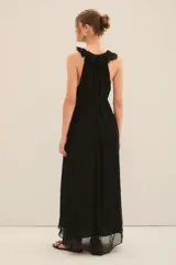 Vestido largo tipo musculosa con escote profundo en el frente. Confeccionado en gasa con forrería a tono. Posee corte en la cintura acompañado de un cinturón de la misma materialidad. Tiene un volado en gasa que se extiende por toda la abertura del cuello. Posee lazos en el cuello, que permiten atar la prenda con plumas metálicas en su terminación.