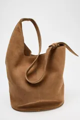 Bolso tipo saca asimétrica de serraje color marrón. Tiene asa bandolera ajustable y cierre con imán. Incluye cartera con bolsillo extraíble.