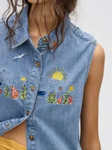 Camisa sin mangas de denim liviano color camel, con cuello camisero y botones frontales. Presenta bordados temáticos marinos en el pecho, incluyendo un sol, olas, una gaviota y vida marina.