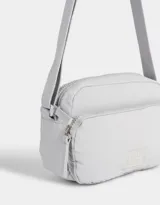 Bandolera gris claro de tela deportiva con bolsillo frontal, correa larga regulable, acceso con cierre superior y tag de goma.