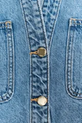 Camisa de denim celeste de corte crop, con cuello solapa, escote en pico, manga corta y dos bolsillos delanteros con botón. Cierre frontal con botones metálicos.