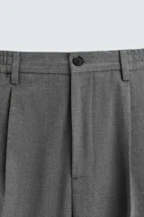 Pantalón negro de corte ancho con cintura elástica, pliegues frontales, bolsillos laterales y traseros de vivo. Tiene detalle de hebilla metálica en la trabilla y cierre frontal con cremallera y botón.