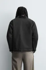 Sobrecamisa de denim de algodón negro con efecto lavado, corte relajado (relaxed fit). Presenta cuello solapa, cierre frontal con cremallera, manga larga con puño abotonado y bolsillo plastrón en el pecho.