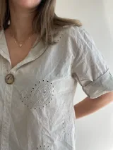 Blusa color crema con cuello camisero, mangas cortas con dobladillo y cierre frontal con un botón de madera. Presenta bordados con motivos de paisley y perforaciones.