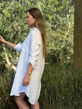 Vestido camisero blanco de lino, con cuello camisero, manga larga con botón para recoger, cierre frontal con botones de nácar, bolsillo en el pecho y diseño asimétrico más largo en la parte trasera.