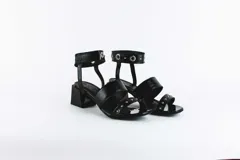 Sandalias negras de cuero con dos tiras sobre el empeine y pulsera al tobillo con ojalillos y tachas. Taco cuadrado de 5 cm.
