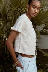 Chaqueta de punto color crema con cuello redondo, manga corta con volantes, bolsillos de plastrón y cierre frontal con botones metálicos.