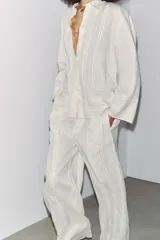 Camisa de lino color crudo, con cuello solapa y escote en pico. Tiene mangas por debajo del codo y detalle de entredós combinados a tono. El bajo es asimétrico y el cierre frontal es con botones.